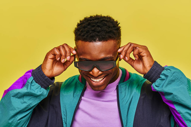 стильный африканский мужчина в красочной куртке регулируя солнцезащитные очки и улыбаясь на желтом фоне - Фото, изображение