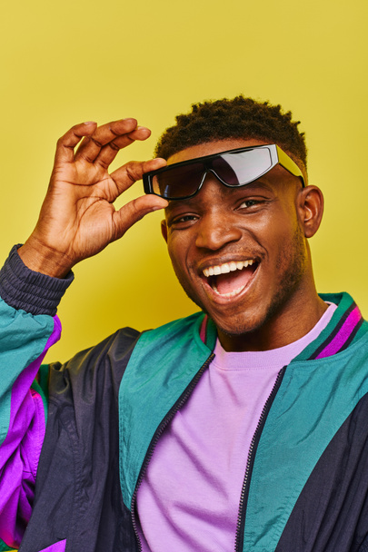 Unbekümmerter afrikanisch-amerikanischer Typ mit trendiger Sonnenbrille und farbenfroher Jacke blickt in die Kamera auf gelb - Foto, Bild