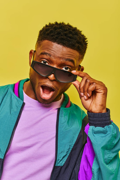 впечатленный африканский американец с открытым ртом, держащий солнцезащитные очки и смотрящий на камеру на желтом - Фото, изображение