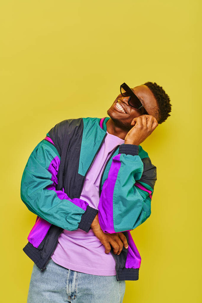 cheerful stylish african american guy in bright windbreaker jacket and sunglasses smiling on yellow - Photo, Image