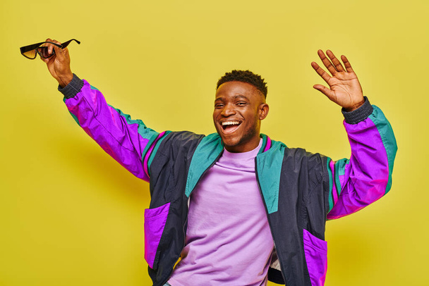 excited african american man in bright jacket holding sunglasses and dancing on yellow backdrop - Zdjęcie, obraz