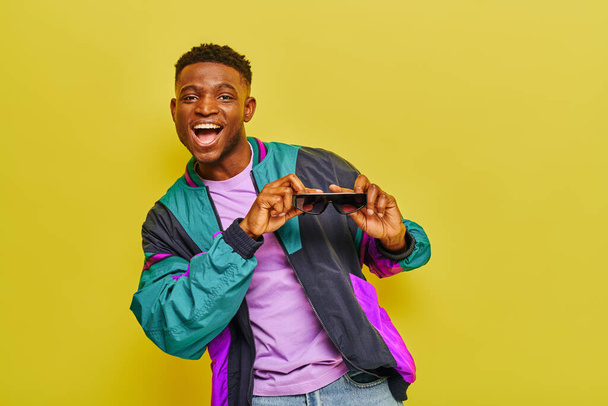 overjoyed african american man in bright jacket holding sunglasses and dancing on yellow backdrop - Photo, Image
