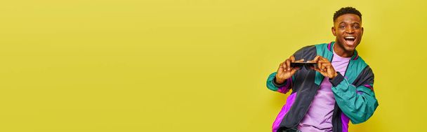 χαμογελαστός Αφροαμερικάνος με φωτεινό σακάκι που κρατάει γυαλιά ηλίου και χορεύει σε κίτρινο, λάβαρο - Φωτογραφία, εικόνα