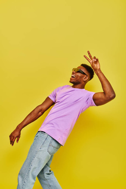gai afro-américain gars en lunettes de soleil et t-shirt violet montrant signe de victoire sur jaune - Photo, image
