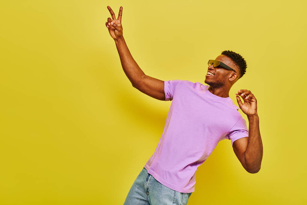 χαμογελαστός Αφροαμερικάνος με κομψά γυαλιά ηλίου και βιολετί t-shirt που δείχνει το σήμα της νίκης στο κίτρινο - Φωτογραφία, εικόνα