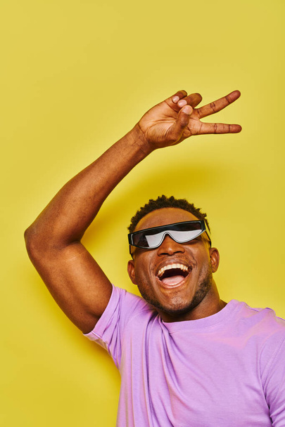 ravi homme afro-américain élégant dans des lunettes de soleil montrant signe de victoire et crier sur jaune - Photo, image