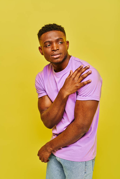 młody przystojny Afroamerykanin mężczyzna w fioletowy t-shirt pozowanie i patrząc z dala na żółtym tle - Zdjęcie, obraz