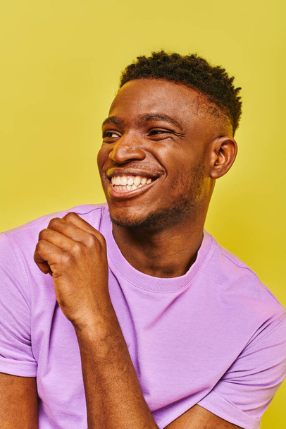 homme afro-américain insouciant en t-shirt violet souriant et regardant loin sur fond jaune - Photo, image