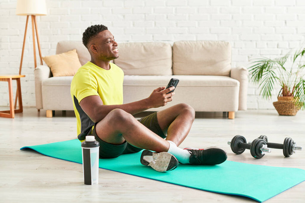 счастливый африканский американец в спортивной одежде сидит на фитнес-коврик со смартфоном рядом с гантелями - Фото, изображение