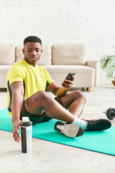 gelukkig Afrikaans amerikaanse man in sportkleding zittend op fitness mat met smartphone in de buurt halters - Foto, afbeelding