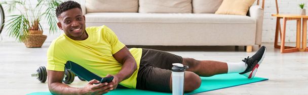 glimlachende zwarte man met smartphone op zoek naar camera op fitness mat in de buurt van sportfles, banner - Foto, afbeelding