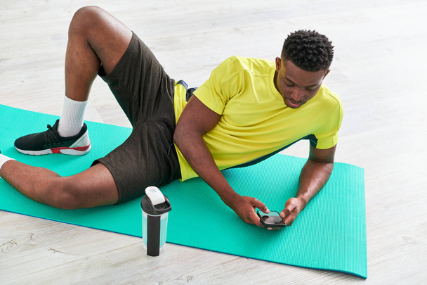 νεαρός αθλητικός αφρικανός Αμερικανός άνθρωπος μηνυμάτων στο smartphone για fitness mat κοντά σε αθλητικό μπουκάλι - Φωτογραφία, εικόνα