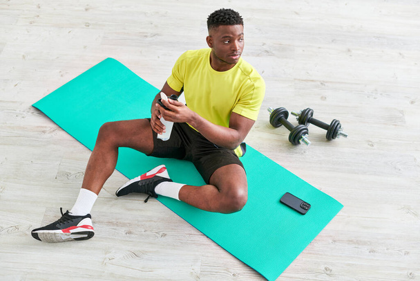 mladý africký Američan chlap ve sportovním oblečení sedí na fitness podložka se sportovní láhve v blízkosti smartphone - Fotografie, Obrázek