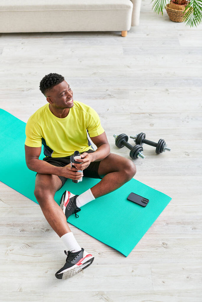 Χαρούμενος Αφροαμερικάνος άνδρας σε αθλητικό ντύσιμο κάθεται σε στρώμα γυμναστικής με αθλητικό μπουκάλι κοντά στο smartphone - Φωτογραφία, εικόνα