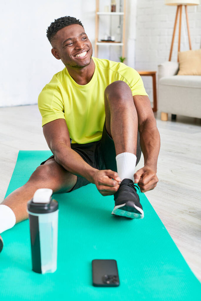 chico afroamericano positivo atando zapatilla de deporte en la estera de fitness cerca de teléfono inteligente y botella de deportes - Foto, imagen