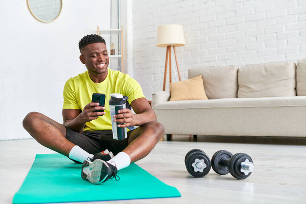 vreugdevolle Afrikaanse Amerikaanse man met sportfles browsen sociale media op smartphone op fitness mat - Foto, afbeelding