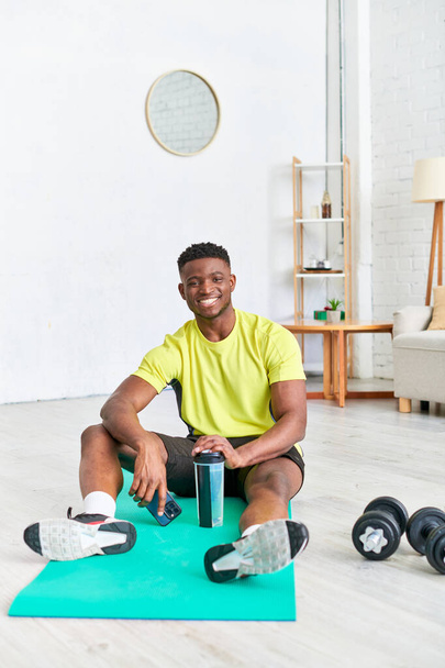 carefree african american man with sports bottle and mobile phone looking at camera on fitness mat - 写真・画像