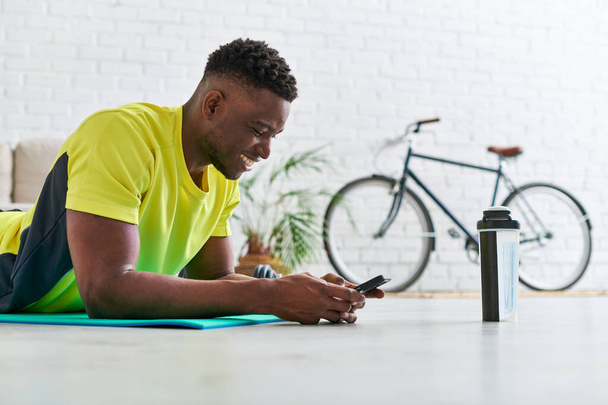 gioioso uomo africano americano sportivo con smartphone sdraiato sul tappeto fitness vicino alla bottiglia sportiva - Foto, immagini