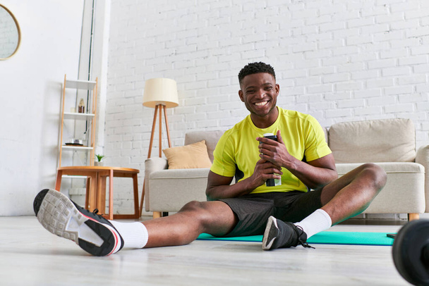 vreugdevolle Afro-Amerikaanse man in stijlvolle sportkleding zittend op fitnessmat met sportfles - Foto, afbeelding