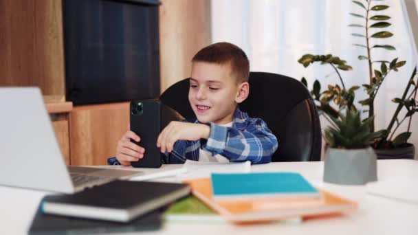 Relaxed caucasian kid in cozy outfit playing games on cell phone in from of laptop and colorful copybooks at home. Elementary pupil getting distracted from remote studying and using gadgets for fun. - Footage, Video