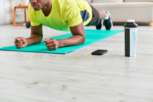 beschnitten afrikanisch-amerikanischer Mann trainiert in Plank-Pose auf Fitnessmatte in der Nähe von Sportflasche und Smartphone - Foto, Bild