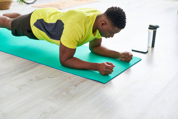 sportlich afrikanisch-amerikanischer Typ beim Training in Plankenpose auf Fitnessmatte in der Nähe von Sportflasche und Smartphone - Foto, Bild