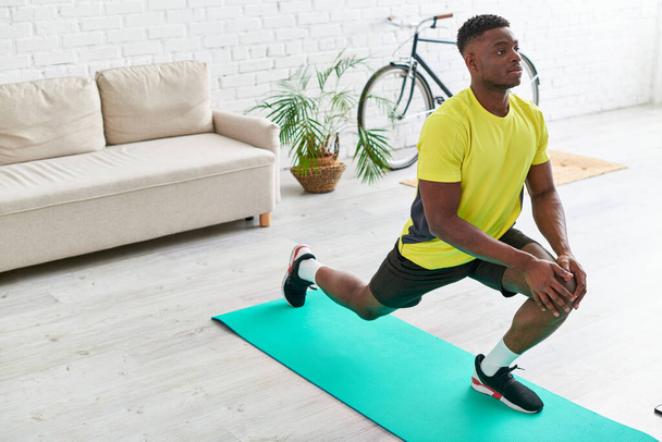 handsome african american man in sportswear stretching on fitness mat in living room at home - Foto, Imagem