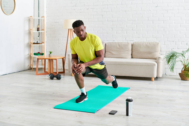 chico afroamericano deportivo haciendo ejercicio en la colchoneta de fitness cerca de la botella de deportes y el teléfono inteligente en casa - Foto, imagen
