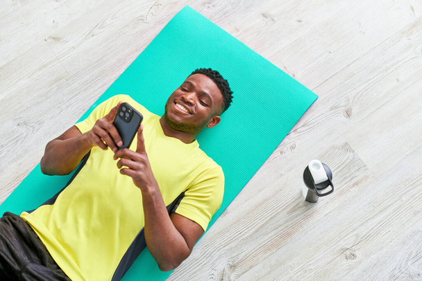cheerful african american man lying down on fitness mat with smartphone near sports bottle, top view - Photo, Image