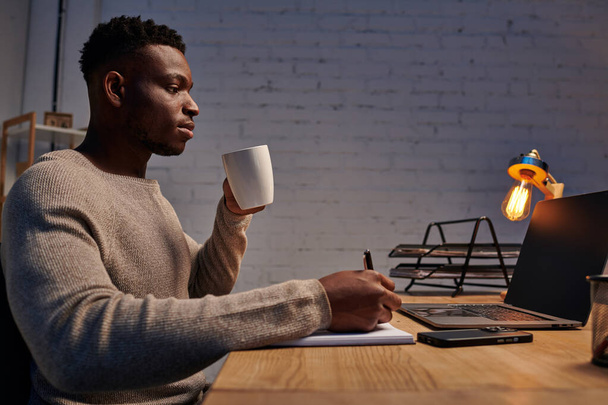 afroamericano chico con taza de café escribir en portátil cerca de portátil de trabajo en casa oficina por la noche - Foto, imagen