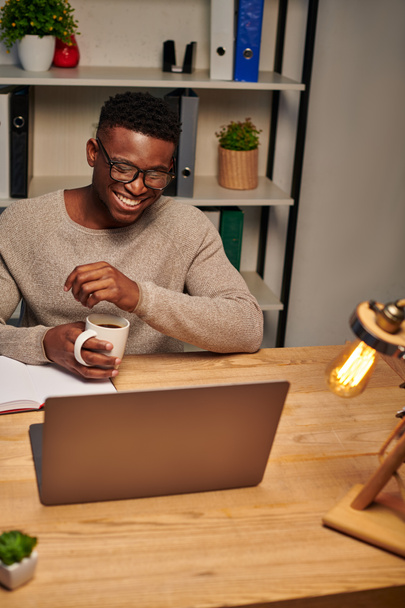 веселий афроамериканський чоловік з чашкою кави сміється під час відеодзвінка в домашньому офісі, фрілансер - Фото, зображення