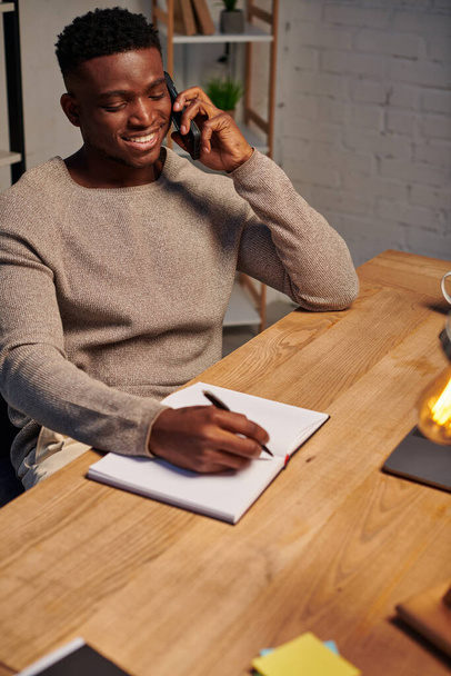 hombre americano africano alegre que habla en el teléfono inteligente cerca del ordenador portátil y del cuaderno mientras que trabaja de hogar - Foto, imagen