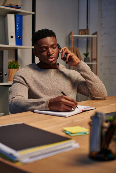 freelancer afro-americano sério falando em smartphone e escrevendo em notebook, trabalho em casa - Foto, Imagem