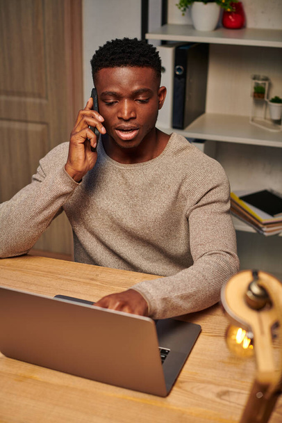 mladý zaměřený africký americký freelancer pracuje z domova a mluví na smartphone v blízkosti notebooku - Fotografie, Obrázek