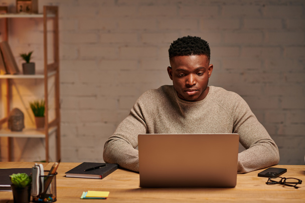 νεαρός σοβαρός αφρικανός Αμερικανός ελεύθερος επαγγελματίας δακτυλογράφηση σε φορητό υπολογιστή στο γραφείο στο σπίτι τη νύχτα, εξ αποστάσεως εργασία - Φωτογραφία, εικόνα