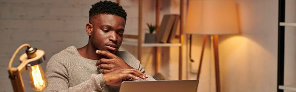δημιουργική αφρικάνικη αμερικανική freelancer σκέψης κοντά φορητό υπολογιστή, ενώ εργάζονται από το σπίτι τη νύχτα, πανό - Φωτογραφία, εικόνα