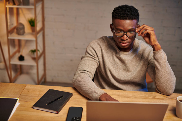 grave afro-américain freelance dans les lunettes de vue penser près d'un ordinateur portable de travail de la maison la nuit - Photo, image
