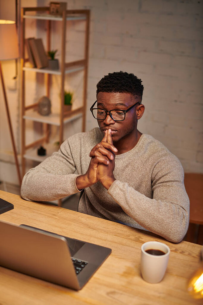 молодий афроамериканський фрілансер в окулярах думає біля ноутбука, що працює з дому вночі - Фото, зображення