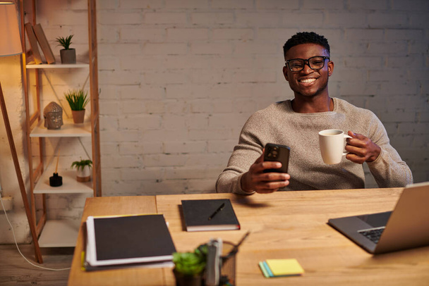 χαρούμενη αφροαμερικανός ελεύθερος επαγγελματίας με καφέ και smartphone κοντά στο laptop τη νύχτα στο γραφείο στο σπίτι - Φωτογραφία, εικόνα