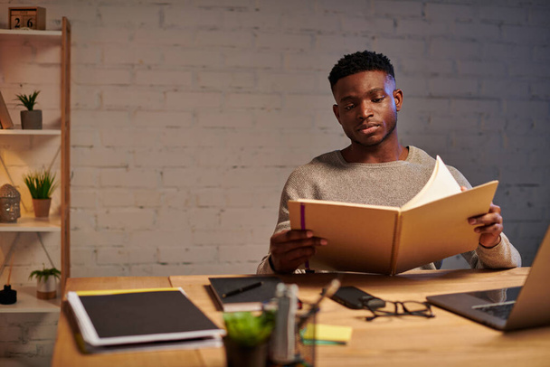νεαρός στοχαστικός Αφροαμερικάνος ελεύθερος επαγγελματίας κοιτάζοντας σημειωματάριο, ενώ εργάζονται στο σπίτι τη νύχτα - Φωτογραφία, εικόνα