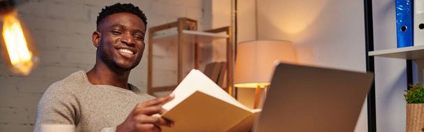 ευχαριστημένος αφρικανός Αμερικανός ελεύθερος επαγγελματίας με σημειωματάριο κοιτάζοντας το φορητό υπολογιστή στο γραφείο στο σπίτι τη νύχτα, πανό - Φωτογραφία, εικόνα
