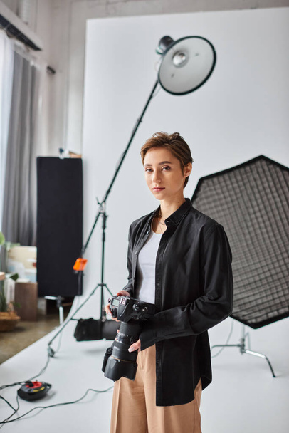 ελκυστική γυναίκα φωτογράφος με κοντά μαλλιά σε casual ενδυμασία κοιτάζοντας την κάμερα, ενώ στο στούντιο της - Φωτογραφία, εικόνα