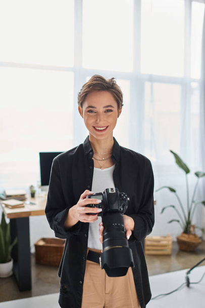 χαρούμενη γυναίκα φωτογράφος με κοντά μαλλιά σε casual ενδυμασία ποζάροντας με κάμερα και χαμογελώντας χαρούμενα - Φωτογραφία, εικόνα
