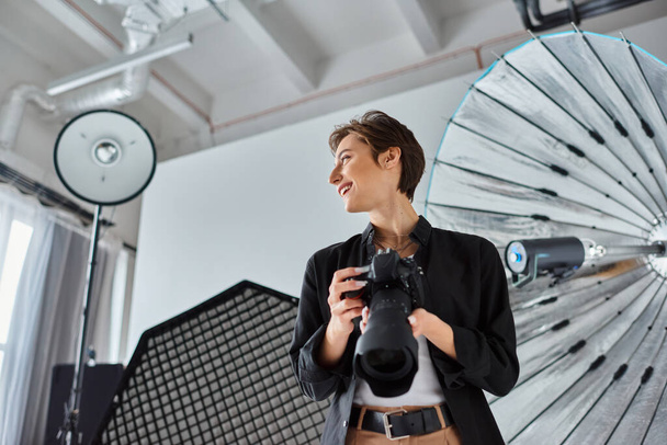veselá žena fotograf v ležérní oblečení s fotoaparátem v ruce s úsměvem a odvrací pohled - Fotografie, Obrázek