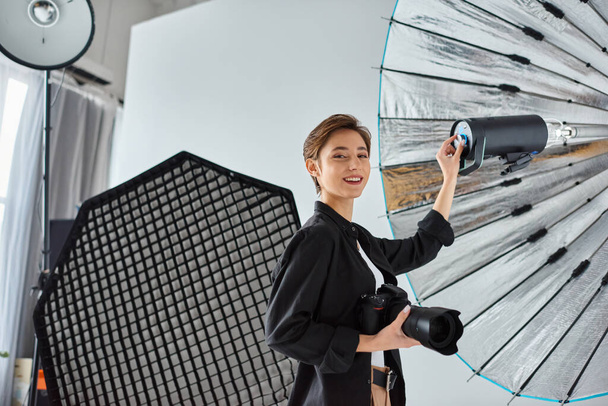счастливая молодая женщина с короткими волосами в повседневной одежде позирует в своей студии с камерой в руках - Фото, изображение
