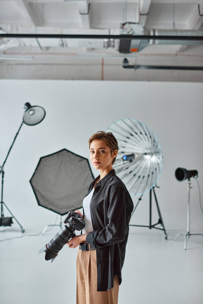 ελκυστική γυναίκα φωτογράφος σε casual ενδυμασία κοιτάζοντας κάμερα που περιβάλλεται από τον εξοπλισμό της - Φωτογραφία, εικόνα