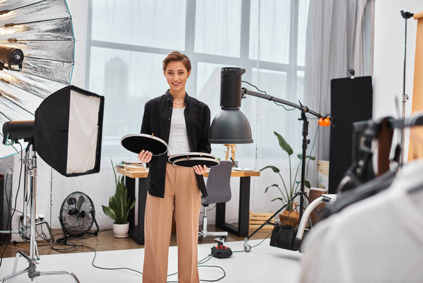 mulher atraente alegre com cabelo curto posando com seu equipamento de fotografia em seu estúdio - Foto, Imagem