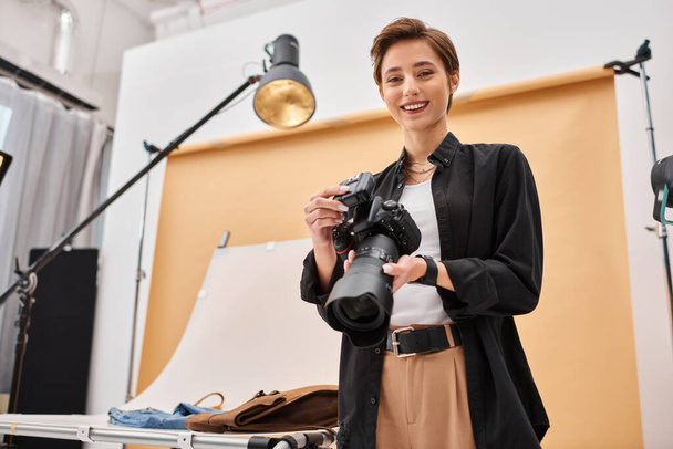 alegre mujer atractiva con equipo de fotografía sonriendo felizmente a la cámara en su estudio - Foto, imagen