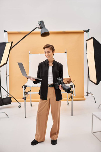 happy attractive woman smiling joyfully while retouching photos and holding her camera in studio - Photo, Image