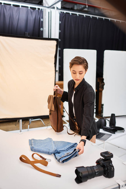 okouzlující krátkosrstá fotografka připravuje fotografie džín a hnědý batoh - Fotografie, Obrázek
